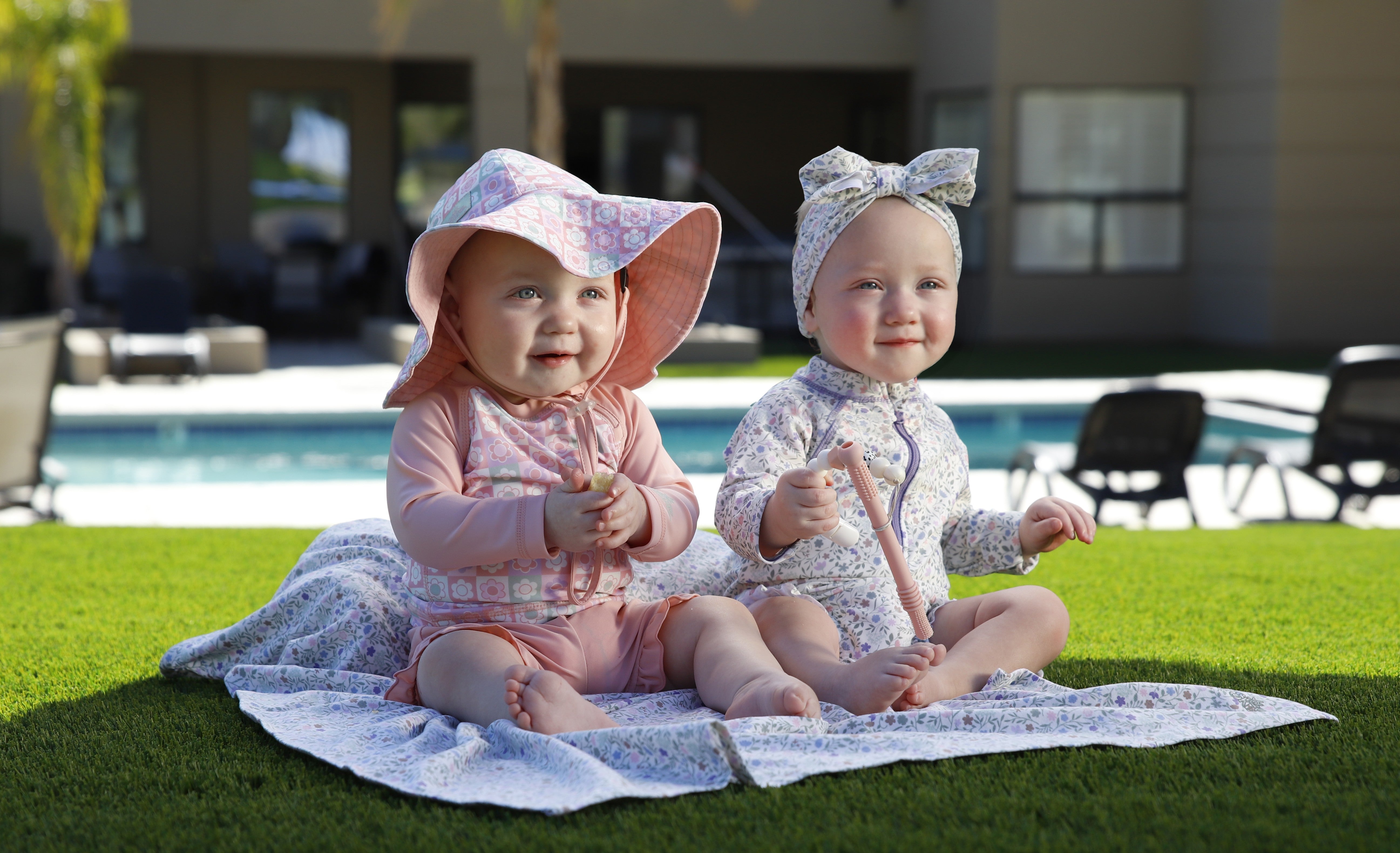 Two babies wearing UV Skinz UPF 50+ swimwear playing sun safe on astroturf by a backyard pool.