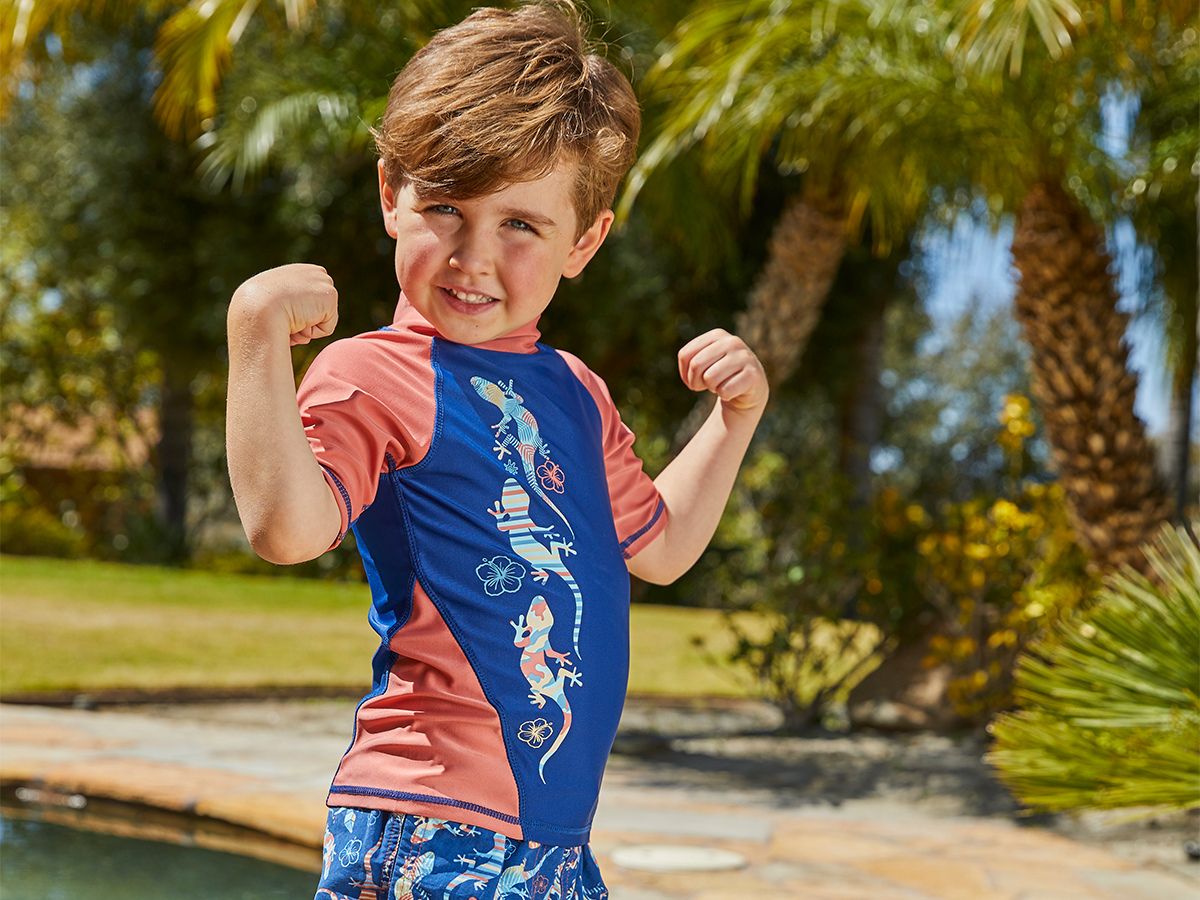 A young boy flexing his arms in a playful pose, standing in front of a lush backyard garden, smiling at the camera.