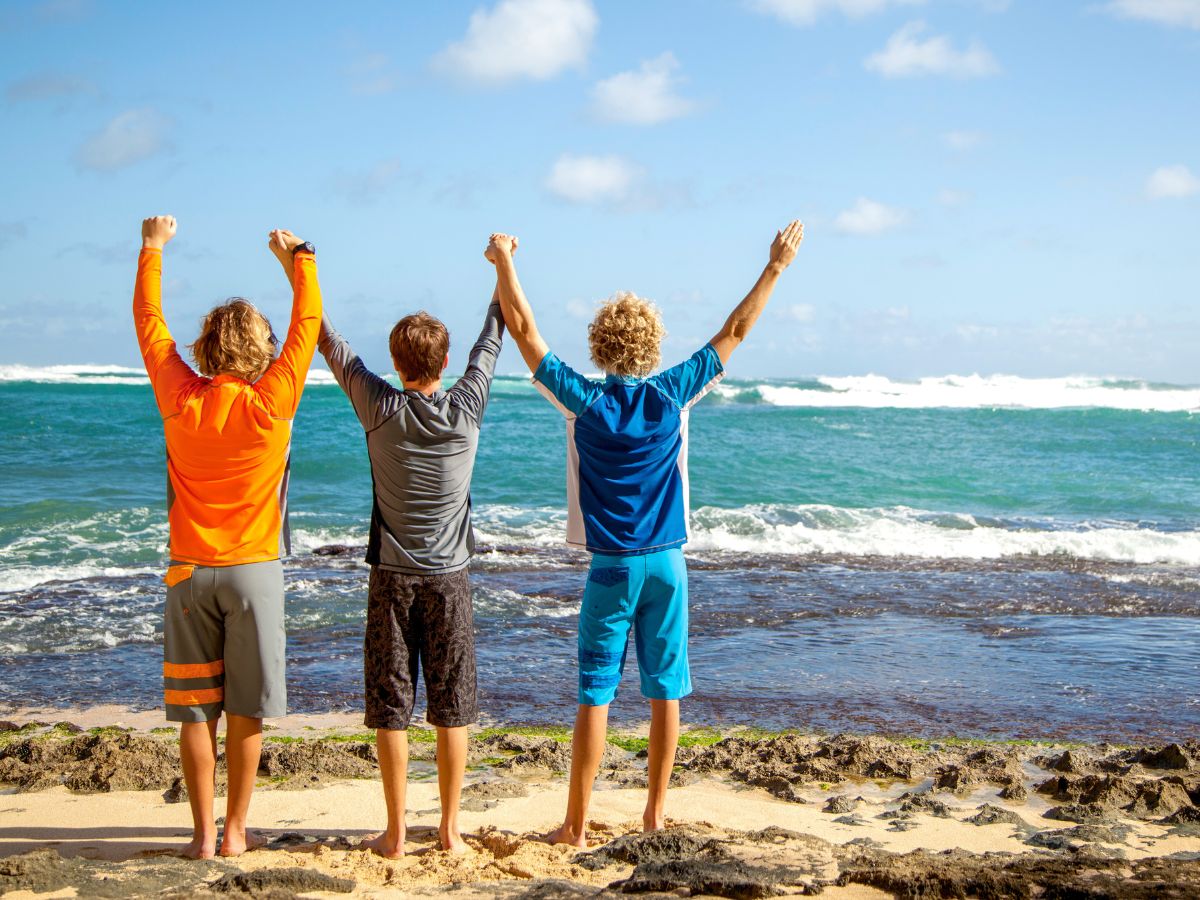 Three boys on the beach wearing full UV Skinz outfits with their hands in the air, celebrating sun safety and staying protected under the sun.