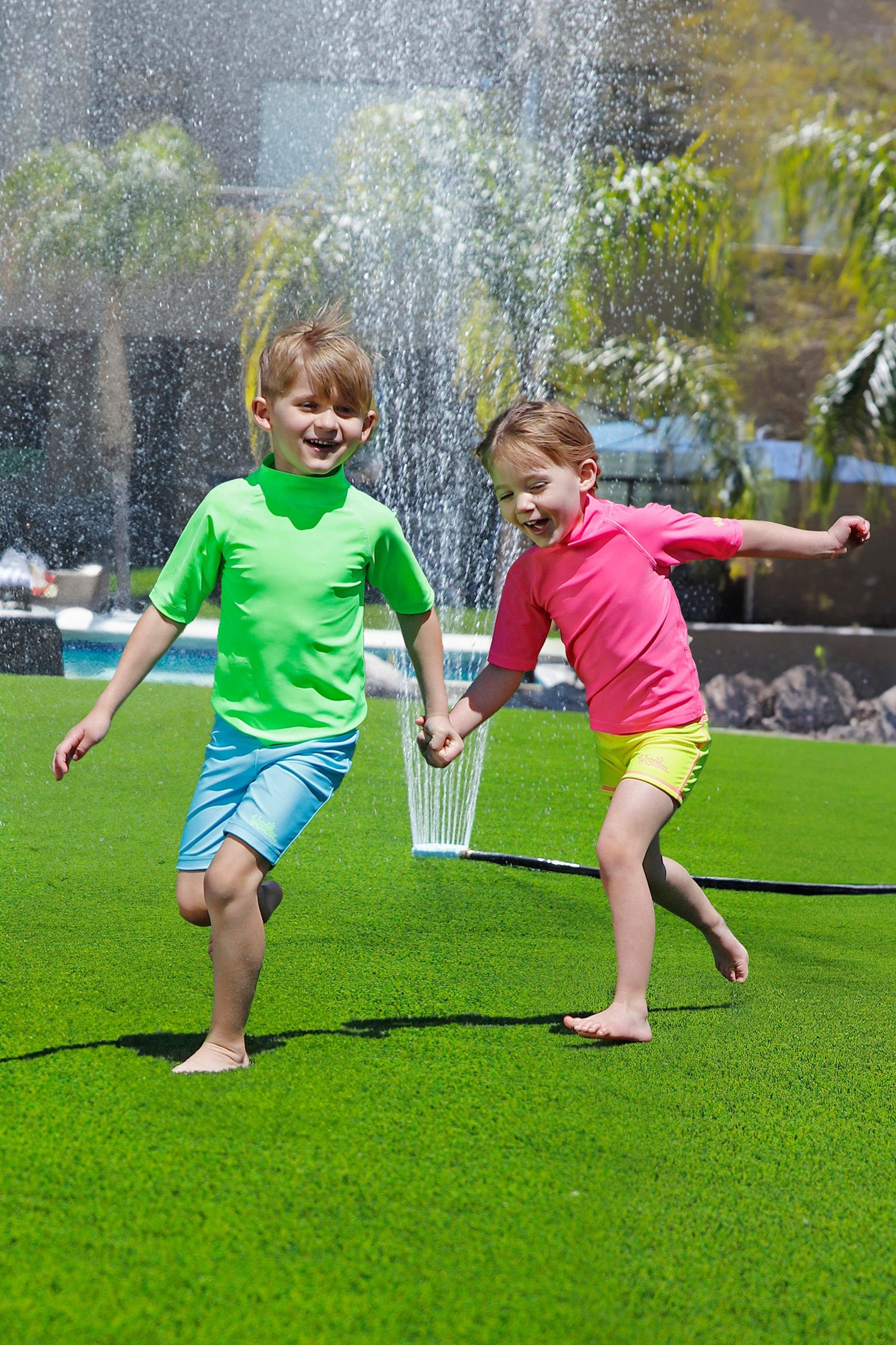 boy having fun in the sprinklers wearing kids neon swim and play jammerz|neon-green
