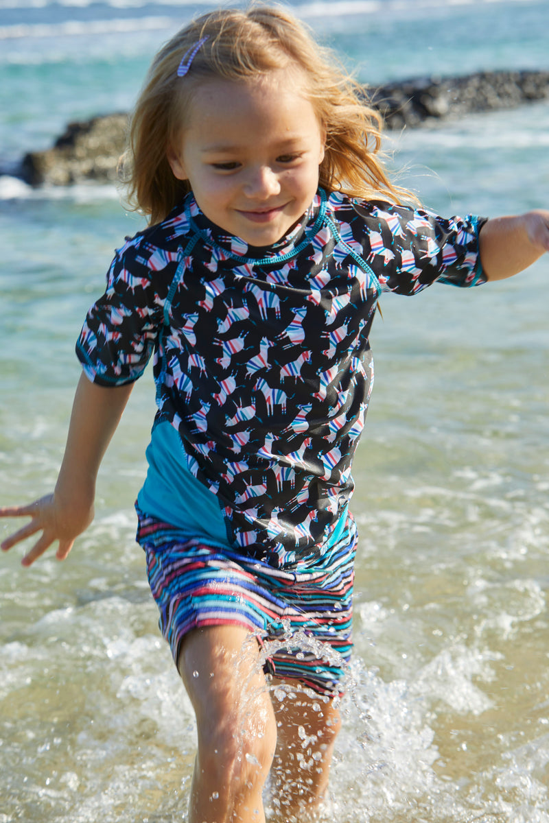 Little Girl Playing in the Ocean in UV Skinz's Girl's Board Shorts in Black Rainbow Stripe|black-rainbow-stripe