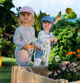 Two babies playing outdoors in a Hawaiian backyard, wearing UV Skinz sun flap hats for sun protection, in front of a vibrant planter box.