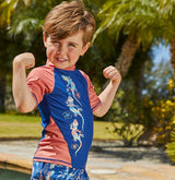 A young boy flexing his arms in a playful pose, standing in front of a lush backyard garden, smiling at the camera.