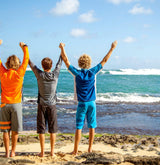 Three boys on the beach wearing full UV Skinz outfits with their hands in the air, celebrating sun safety and staying protected under the sun.